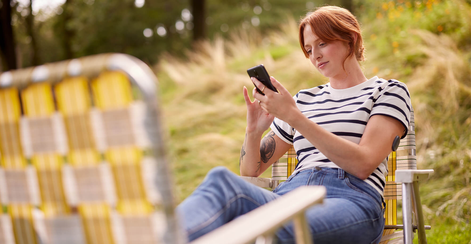 Person reclines in a striped shirt on a yellow lawn chair, using a smartphone outdoors near tall grass.